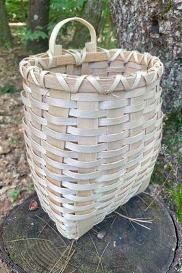 Small handwoven ash basket with a handle displayed on a tree stump in the woods.