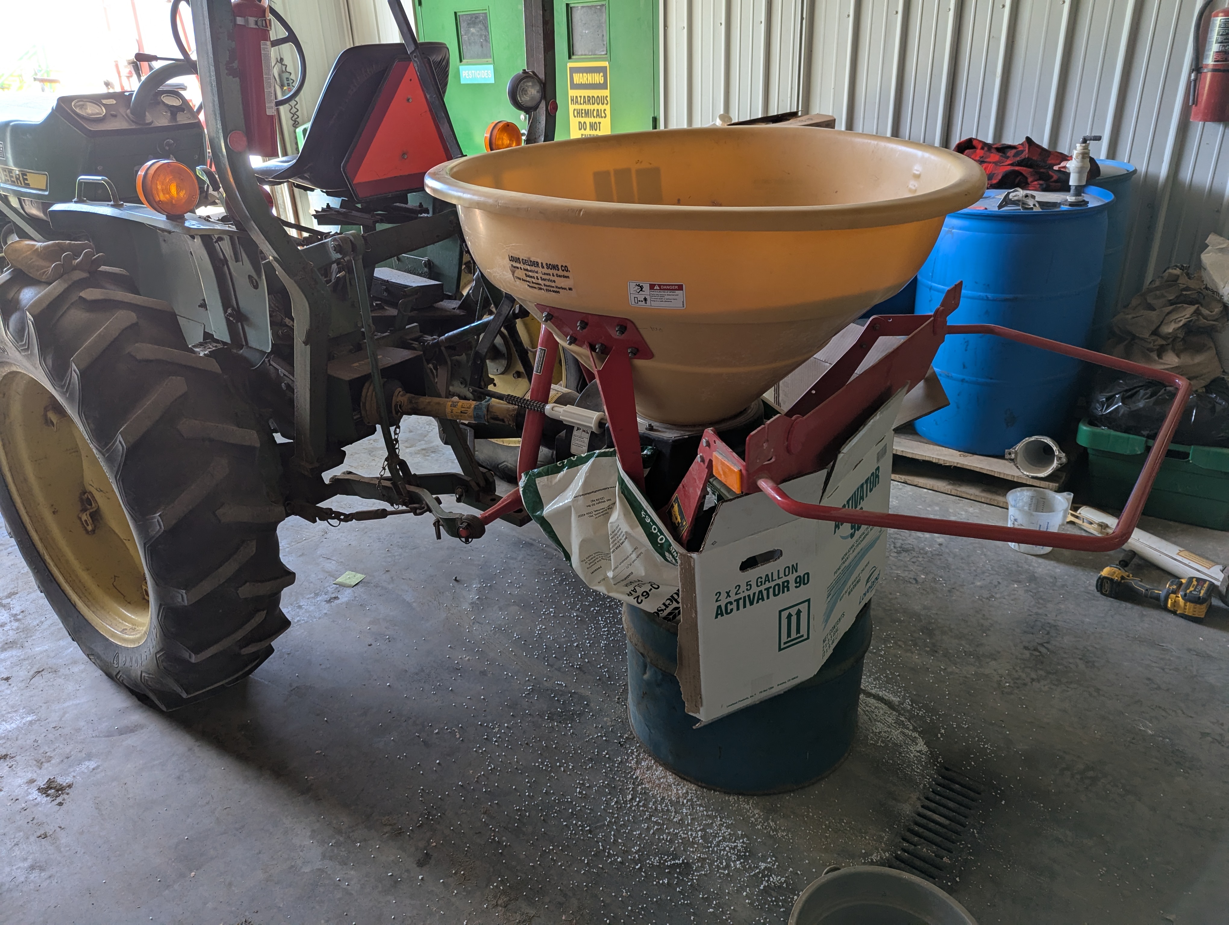 A tractor-mounted broadcast spreader positioned indoors on top of a blue barrel for calibration, with fertilizer pellets scattered on the floor.