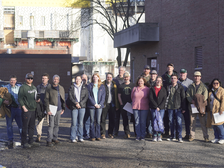 A large group of people stands outdoors in front of industrial buildings for a group photo. The group includes men and women in casual and work attire, some holding papers, with sunlight casting long shadows across the pavement.