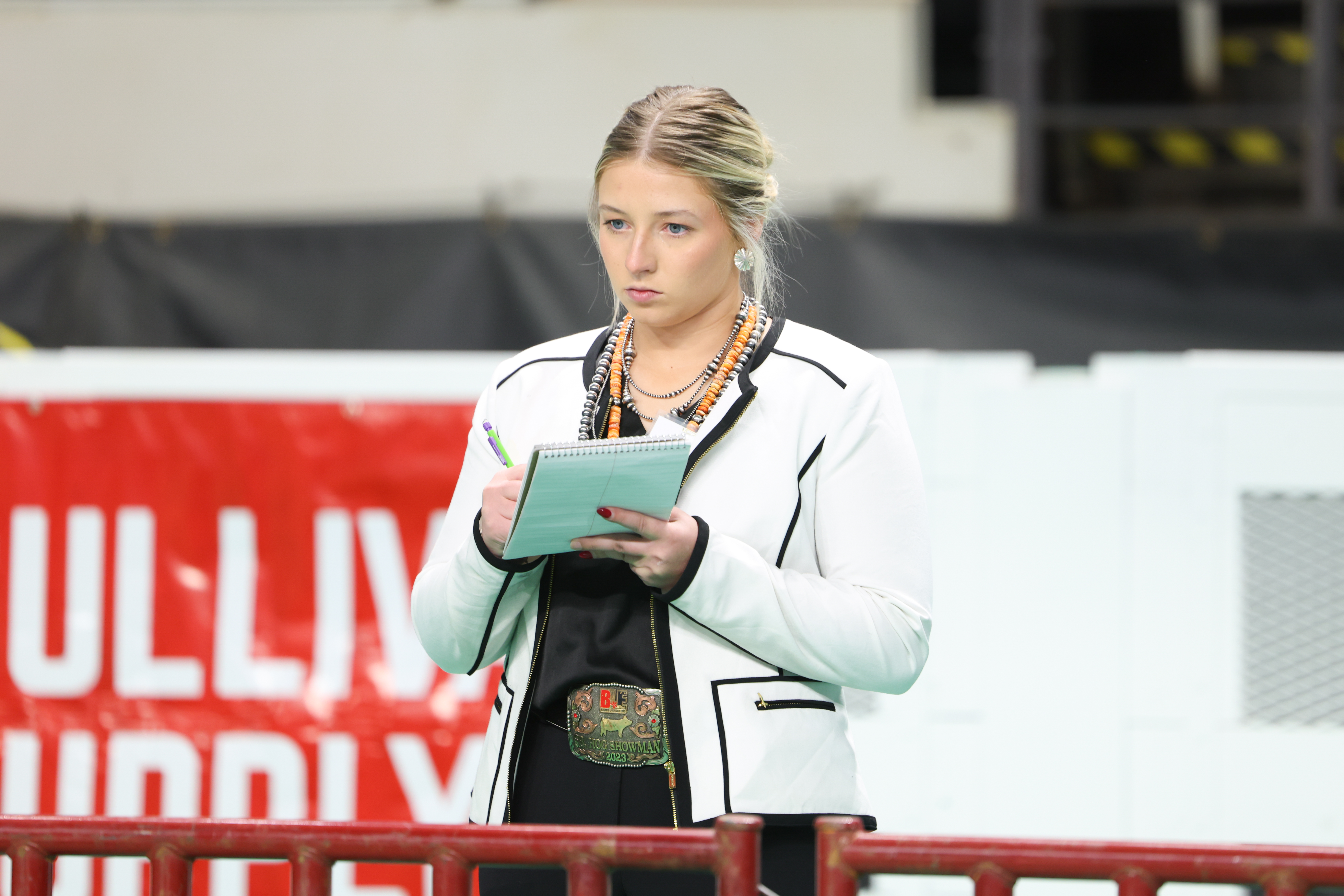 Joscelyn Layman takes notes during a livestock judging event, standing behind a rail and holding a notepad and pen
