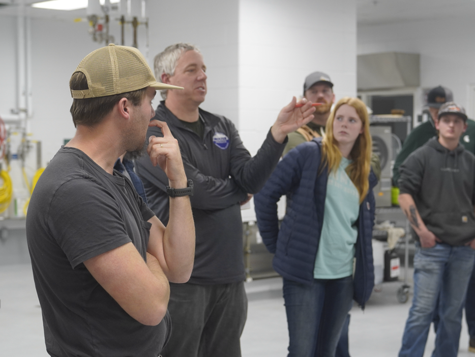 A group of people stands in a lab or processing facility while a man gestures and explains something to them. Others listen attentively, including a woman in a blue jacket and several men in casual workwear. Industrial equipment and pipes are visible in the background.