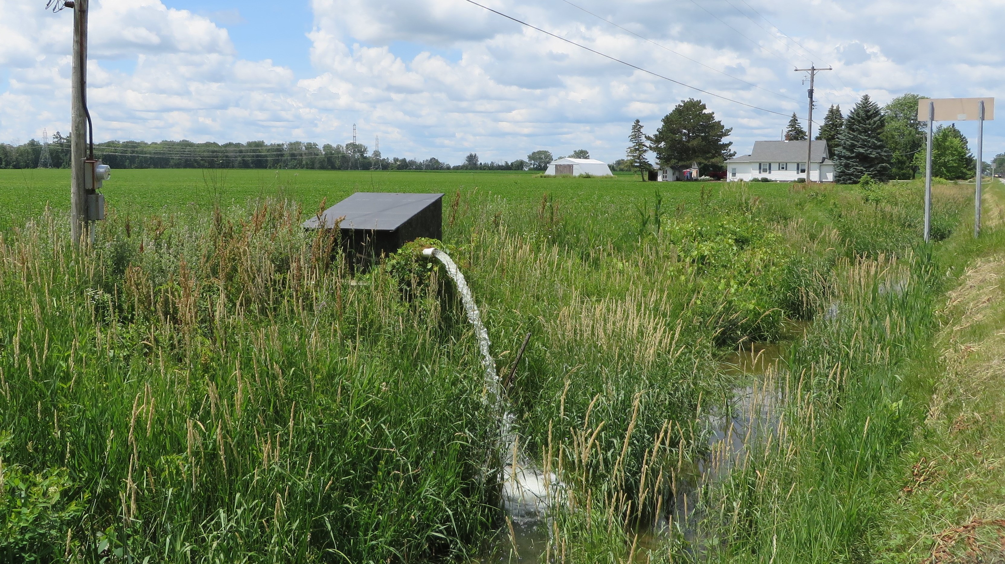 Photo of a nearly level field with not enough grade to allow gravity flow of the drainage water into a shallow drainage ditch, so the drainage water flows into a sump from which it is pumped out to the ditch.