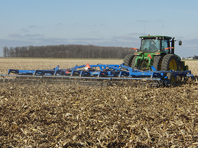 A green tractor pulls a blue Landoll 7450 implement across a corn field during fall tillage, cutting and incorporating crop residue into the soil.